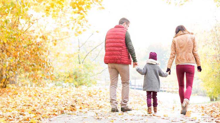 Parents holding their kid's hand walking down a leafy lane in the fall.