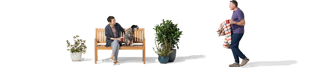 Between a pair of potted plants, a woman sits on a wood bench with her dog. She watches as her husband approaches with a Christmas-colored blanket and a red mug.