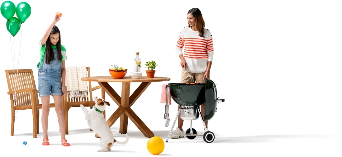 As mom tends the grill next to the outdoor patio furniture, her daughter plays fetch with their Jack Russell terrier A trio of green balloons are tied to the wooden picnic chair.