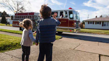 Two young children wave at a passing fire truck from their driveway.