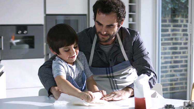 A dad helps his son as they roll out some dough.