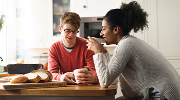 Una pareja comparte una taza de café en su mesa de la cocina. Un pan rebanado está en la tabla de cortar.