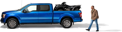 A man approaches his blue pickup truck with a black ATV in the bed.