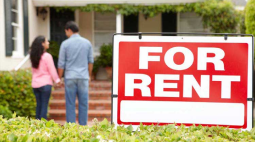 With a For Rent sign in the foreground, a young couiple stand at the bottom of the stairs to a rental home.