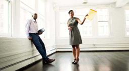 A female landlord with a yellow pad shows off an empty corner apartment while her new tenant leans against the windowsill.