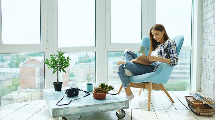 A young woman relaxes with a book in her blue chair next to the window of her new condo.