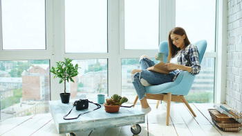 Una joven se relaja con un libro en su silla azul junto a la ventana de su nuevo condominio.